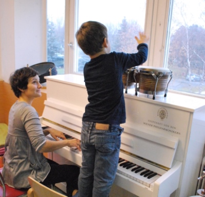 Kleiner Junge spielt mit Trommeln, während eine Musikpädagogin am Klavier spielt